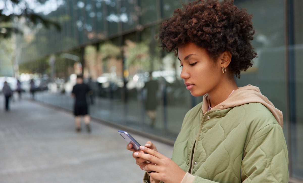 A young Black woman standing on a city sidewalk views search results on her mobile phone