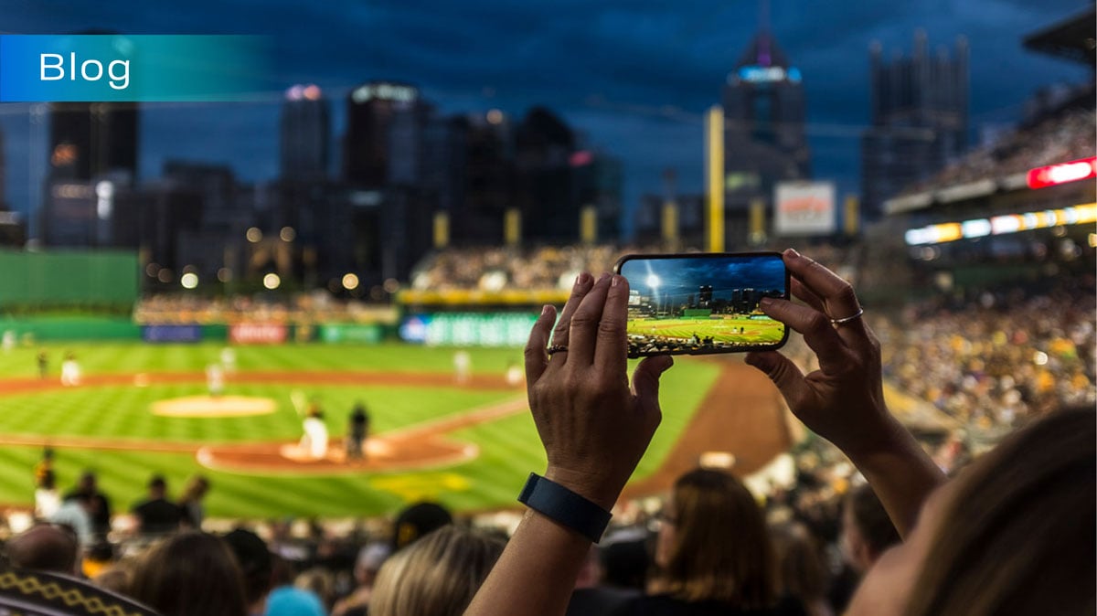 A photo of a spectator using their camera to take a photo at a live baseball game in order to post it to social media. The word 'Blog' is overlaid on the upper-left corner