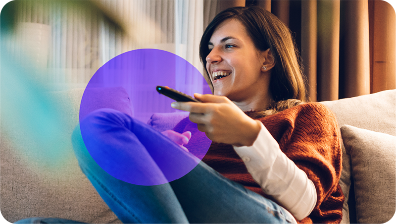 A woman sitting on her sofa points a remote at her smart TV. A purple circle overlaid around the remote draws attention to the device